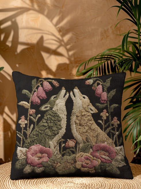 A square woven tapestry pillow with two howling wolves on a black background. The wolves are surrounded by pink and green botanical flowers, creating a vintage, folkloric scene. This decorative cushion is perfect for cottagecore, dark academia, and gothic home decor.